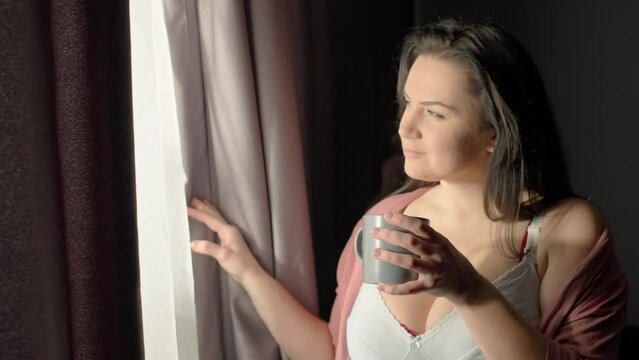 Caucasian woman opens the curtain and drinks morning coffee while looking out the window. Happy girl at home. Concept of warmth and smile. High quality 4k footage