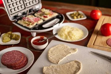 Cooking waffles stuffed with sausage, cheese. Pizza waffle, piffle. Various ingredients on the kitchen table.