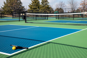 View of a pickleball complex with a paddle and yellow ball on blue and green courts beside a playground in a suburban park in early spring.