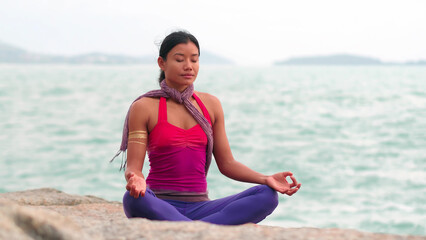 Asian woman meditating in yoga lotus pose on stony sea beach