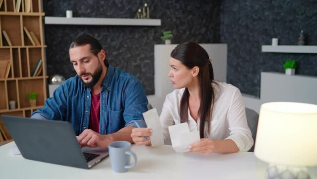 Husband and wife, a young couple sitting at the table sorting out checks bills budget. Young woman holding notes checks man typing on laptop sitting at table at home camera moves left