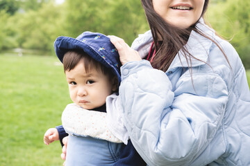 Close-up photo of a male infant carried by his smiley mother on a baby carrier wearing a blue hat walking at the park. He is sad and unwell and looking at camera. Multicultural and multiracial family.