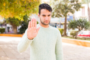 young hispanic man looking serious, stern, displeased and angry showing open palm making stop gesture