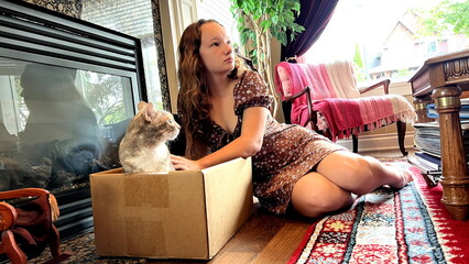 A teenage girl is stroking a gray tabby cat that is in a box near the fireplace on the left side a doll in rocking chairs on the right and a red carpet the room is expensive and decorated.