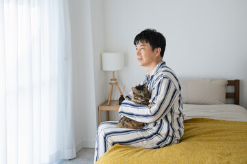 ベッドに座って猫を抱くパジャマ姿の50代男性