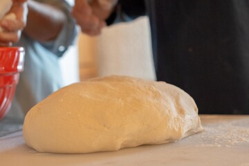 A large bun of dough rises on a white plastic board with a red glass bowl in the background. The large ball of dough is made with white flour. There's a window in the background with natural light.
