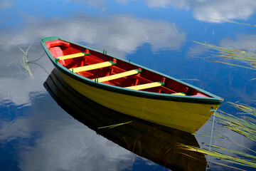 A small yellow wooden row boat with green trim and a red interior floats on calm water with reeds...