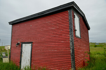 Fototapeta na wymiar The exterior of a colorful vintage red storage building. The old wooden barn has wood siding on the walls, a small window, and a white closed wooden door. There's green trim on the edge of the shed. 