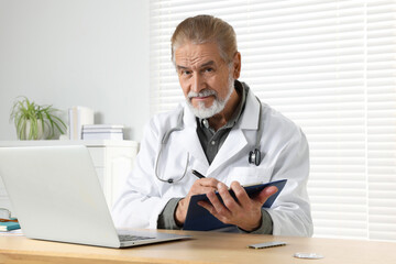 Senior doctor with laptop and clipboard consulting patient at wooden desk in clinic. Online medicine