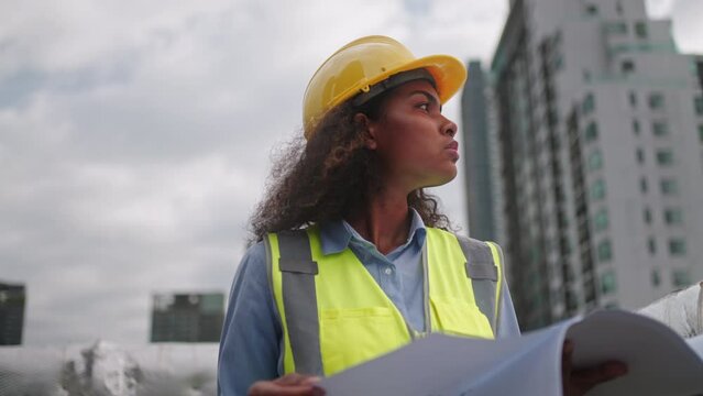 Civil engineer woman dark skin wearing uniform and safety helmet under inspection,checking plan on construction site by paper blueprint.Civil,Industry,construction,building construction engineering