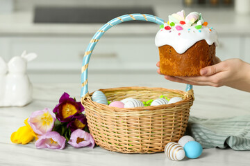 Fototapeta na wymiar Woman holding tasty Easter cake near wicker basket with painted eggs and flowers at white marble table, closeup
