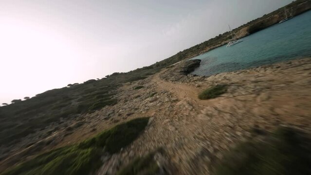 FPV drone fly low over rocky terrain along the seaside with sailboats  in Cala Varques, Mallorca, Spain at daytime.