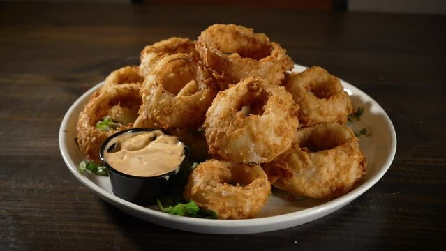 Large deep fried onion rings piled high on white plate with sauce, slider 4K