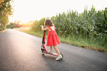 girl in red dress riding red scooter on a road next to corn field