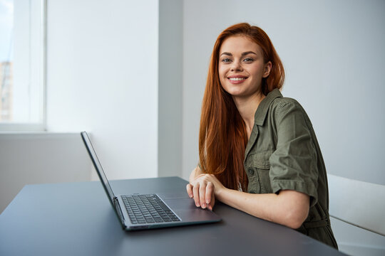portrait of a cute, smiling red-haired woman sitting at a laptop with her hands folded on the table and looking at the camera
