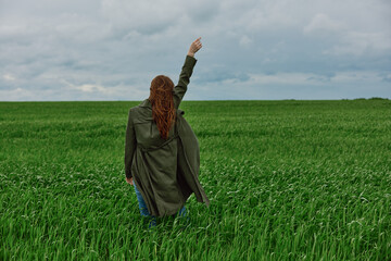 a red-haired woman stands with her back to the camera in a long coat with her hand raised up in a green field in rainy weather