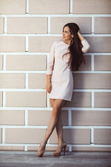 beautiful young woman against white brick wall An attractive girl with long beautiful tanned legs is touching her dark long hair. She is wearing a short white dress and high heels.