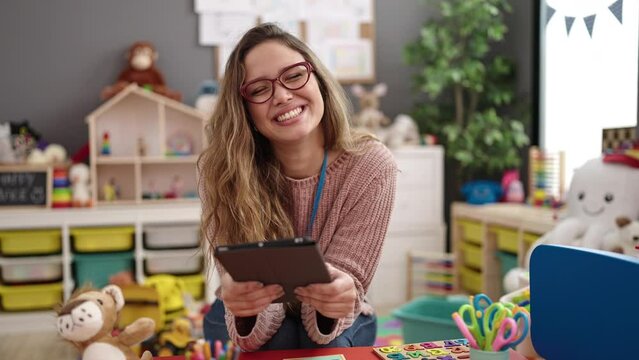 Young beautiful hispanic woman preschool teacher smiling confident using touchpad at kindergarten