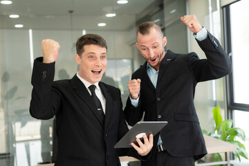 Two handsome young businessmen in suits holding tablet excitedly celebrating success about a real estate project or having a video call against the background of an office building.