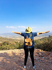 Adult woman with hat looks towards the horizon on top of a mountain opens his arms and breathes in front of the mountainous landscape proud of her achievement