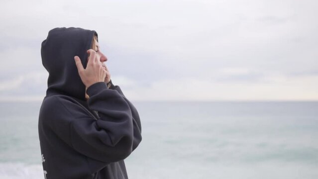 A relaxed woman in a warm sweatshirt with a hood on her head looks out to sea