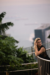 A beautiful Caucasian blonde girl in a luxurious black dress is standing against the backdrop of a park in a modern city. Greenery and concrete in the city of the future. Black dress concept