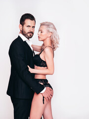 Sexy beautiful woman and her handsome boyfriend. Young passionate couple hugging. Man in elegant black suit. Blond model in lace lingerie wearing pure underwear. Posing in studio, near white wall