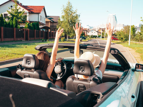 Portrait of two young beautiful and smiling hipster female in convertible car. Sexy carefree women driving. Positive models riding and having fun outdoors. Enjoying summer days. Raising hands