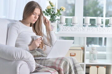 Young woman sitting in armchair with cup of tea and laptop on knees