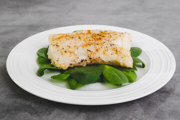 Cod fillet baked with garlic butter sauce served with fresh spinach on a white plate close-up on...