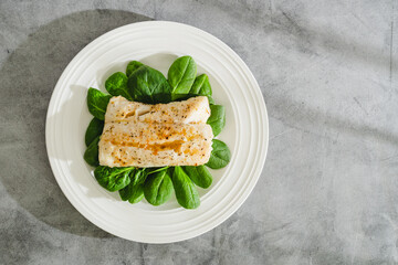 Cod fillet baked with garlic butter sauce served with fresh spinach close-up on a white plate, flat lay