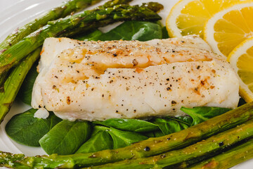 Cod fillet baked with garlic butter sauce served with fresh spinach and roasted asparagus on a white plate close-up