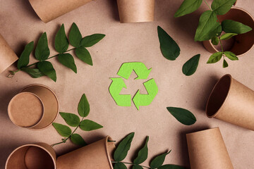 Green arrows recycle symbol with disposable empty paper coffee cups and green leaves on brown paper background.