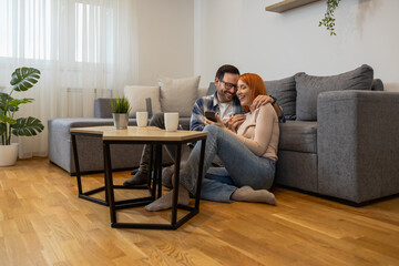 Happy young woman and man using smartphone together, sitting on floor in modern living room