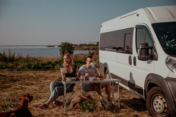 Couple of travelers on van have breakfast in nature against the background of the sea with dog