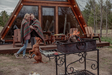 Couple embraces wrapped in blanket on the porch of campsite house next to dog and campfire