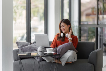 Beautiful Asian Woman smile and relaxing at home and using laptop computer sitting on cozy sofa