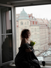 woman with curly hair standing near window, with bouquet of tulips
