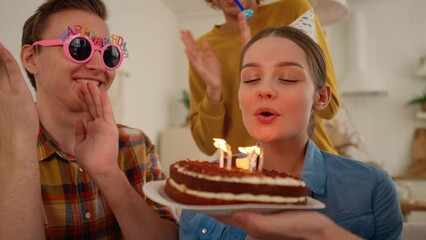 Surprise for birthday girl from friends. Friend closes eyes with hands. She happy about birthday cake with burning candles. Guy in funny glasses blowing party horn. Celebration, festive mood concept.