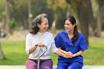 Smiling Asian female nurse holding senior woman hand having pleasant conversation during sitting...