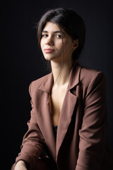 Classic studio portrait of a young brunette dressed in a black top and formal suit, who is sitting on a chair against a black background.