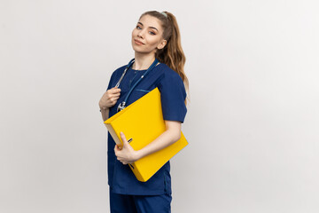 A medical attendant wearing a blue scrubs ensemble holding paperwork against a white backdrop