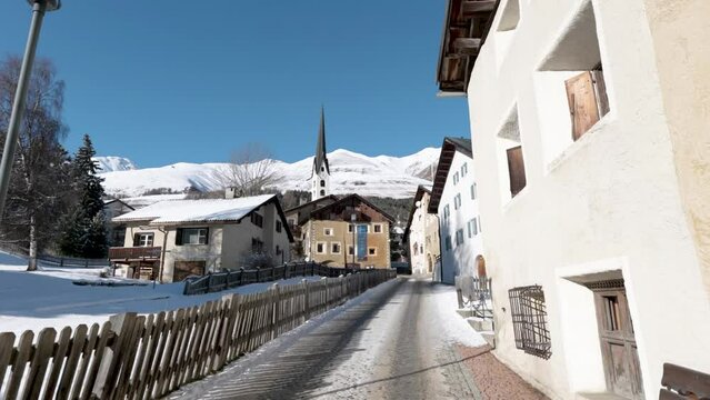 View of a traditional Swiss village streets and houses with mountains in background during a sunny winter day in Zuoz, Switzerland.