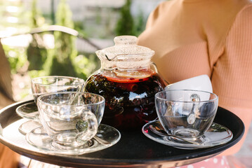 The waitress holds three glass cups and a glass teapot on a tray. Tea items close-up. Sunny day, street cafe