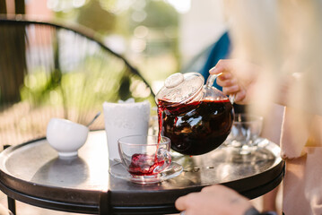 In a street cafe, in the morning, a waiter pours strong tea from a transparent glass teapot into a glass cup. There is a sugar bowl on the table. Blurred background and foreground.