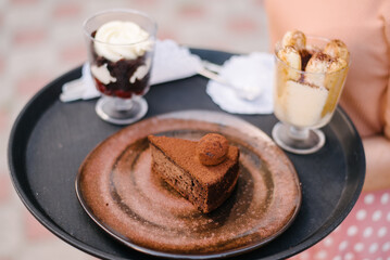 The waiter of a street cafe carries a chocolate cake on a tray on a beautiful chocolate-colored plate, and two glasses with desserts. Chocolate cake in focus. Blurred background with desserts