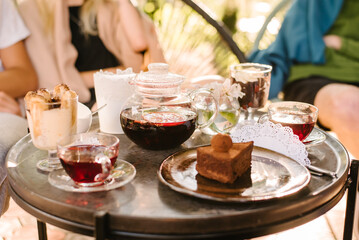 terrace of a summer cafe. on the table are glass cups with tea, a glass teapot with fruit tea and desserts. People are sitting on a blurred background. Focus on the teapot