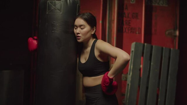 Portrait of tired out of breath attractive athletic fit Asian female kickboxer in boxing gloves embracing punching bag, taking a break and feeling exhausted while working out martial arts training in
