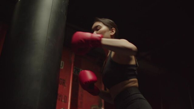 Portrait of determined active sporty fit pretty Asian woman boxer in gloves practicing boxing training, working out and improving jab and uppercut punch techniques with punching bag. in grunge gym.