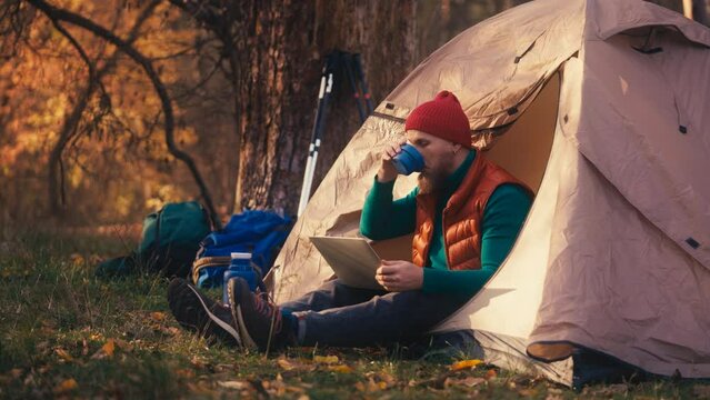 Young man working on laptop by tent in woods, downshifting lifestyle, freelancer